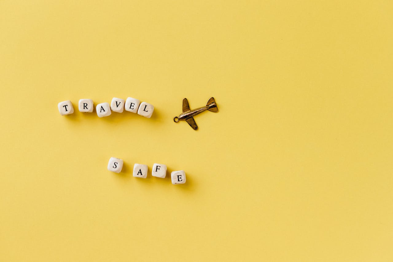 Flat lay of 'Travel Safe' text with airplane pendant on yellow background, evoking travel and safety concepts.